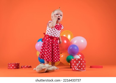 Crying Baby Girl In Red Dress Upset With Birthday Gifts. Studio Photo On Orange Background