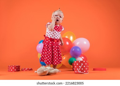 Crying Baby Girl In Red Dress Upset With Birthday Gifts. Studio Photo On Orange Background