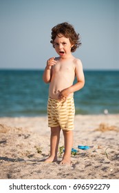 Crying Boy On The Beach. A Small Child Is Standing By The Sea And Crying. The Kid Is Standing On The Sandy Beach.