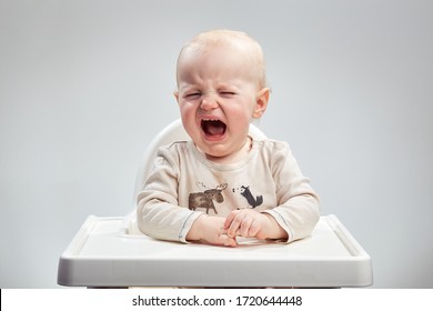 Crying Caucasian Infant Sitting In A High Chair Looking Straight Forward On A Plain White Background.