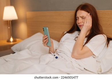 Crying ill red-haired young woman having online video call via smartphone lying in bed at home. Sick redhead female using cellphone to talk with family. Concept of fever, flue or virus.