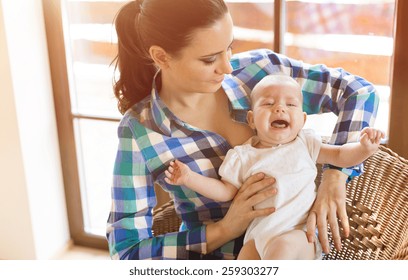 Crying Little Baby In The Arms Of Her Mother In A Living Room.