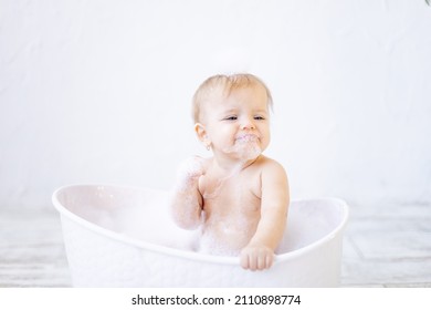 a crying little baby girl with foam in her mouth and in front of her eyes bathes in a bathtub with foam and soap bubbles in a bright bathroom, the concept of child hygiene