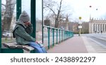 crying lonely child, wiping eyes with hand, in warm cloth at bus stop, blurred street in background. concepts: lost child, sad child waiting on bus stop, crying kid outdoors, child