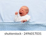 Crying newborn baby boy in bed. New born child sleeping under a blue knitted blanket. Children cry. Bedding for kids. Infant napping in bed. Healthy little kid shortly after birth. Cable knit textile.