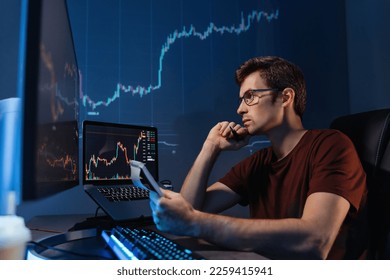 Crypto trader freelancer sitting at table with notepad late night. Noting down info after checking ico and trading market dynamics on his personal computer with candlestick chart on display 