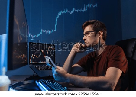 Crypto trader freelancer sitting at table with notepad late night. Noting down info after checking ico and trading market dynamics on his personal computer with candlestick chart on display 
