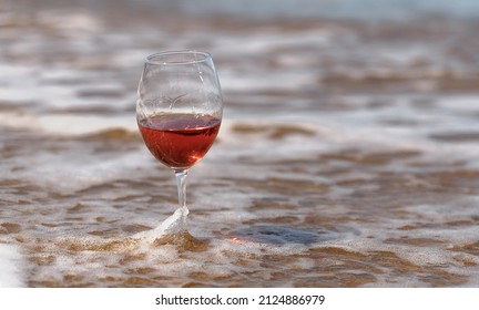 Crystal Glass With Rose Wine Stands In The Sea On The Sand, Against The Background Of A Sea Wave In Defocus