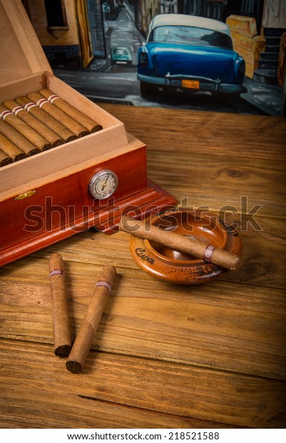 Cuban cigars\
and humidor with ashtray on rustic wooden table with Cuban painting\
of american old car in\
background