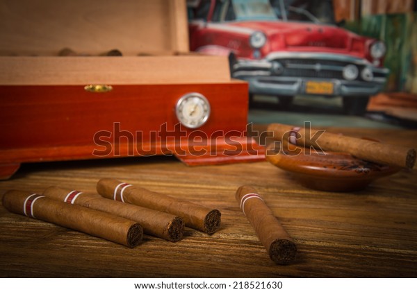 Cuban cigars\
and humidor with ashtray on rustic wooden table with Cuban painting\
of american old car in\
background