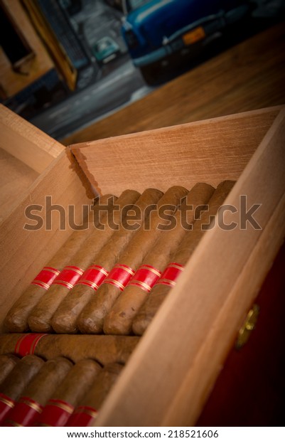 Cuban cigars and humidor\
on rustic wooden table with Cuban painting of american old car in\
background