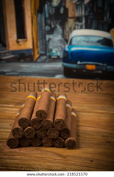Cuban cigars  on rustic wooden table\
with Cuban painting of american old car in\
background