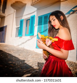 A Cuban girl with long brown hair celebrating her Quinceañera party , posing in a long red dress on the street, holding yellow flowers in her hand. 