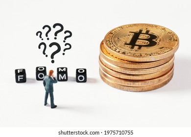 Cubes with FOMO text on white background. Thinking businessman figurine with question marks above his head looking at cubes next to bitcoin stack. Cryptocurrency, blockchain or trading concept.