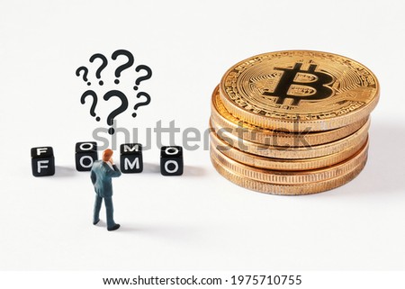 Cubes with FOMO text on white background. Thinking businessman figurine with question marks above his head looking at cubes next to bitcoin stack. Cryptocurrency, blockchain or trading concept.