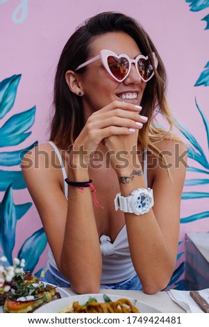 Cucasian happy woman having healthy breakfast in cafe on background of pink tropical wall. Heart trendy sunglasses.