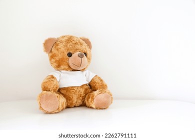 Cuddly brown teddy bear stuffed toy wearing a white t-shirt sitting on white table against white wall, copy space