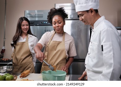 Cuisine course, senior male chef in cook uniform teaches young cooking class students to knead and roll pastry dough, prepare ingredients for bakery foods, fruit pies in stainless steel kitchen.