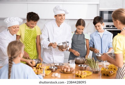 At culinary master class, experienced male chef in white uniform explains to children subtleties of dessert preparation and female assistant helps child to work with whisk - Powered by Shutterstock