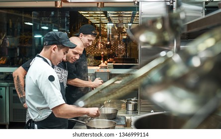 Culinary School. Strict Young Chef In Apron Looking Carefully How His Assistant Cooking At Restaurant Kitchen