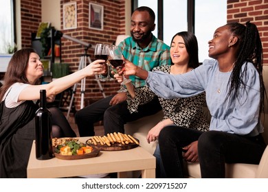 Culturally Diverse Group Of Adults, Men, Women Toasting, Seated At Living Room Couches, Drinking Glasses Of Wine. Ethnically Different People Bunch Enjoying Alcohol, Eating Bread Sticks.