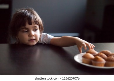 Cunning cute little girl stealing delicious muffin on table, hungry funny impatient child reaching hand to take homemade cookies from plate, unhealthy food and kids sugar craving addiction concept