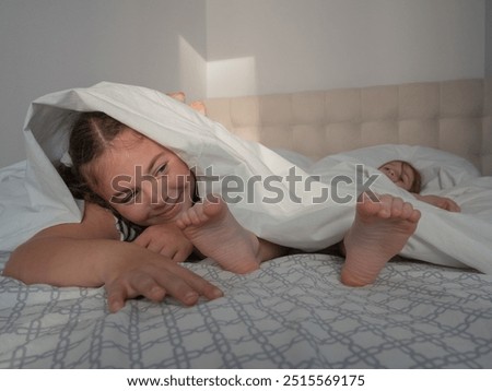 A cunning little girl lies on her stomach on the bed, looks out from under the duvet, next to her sister's bare feet, smiles, pulls her hands to her heels to tickle, laughs.