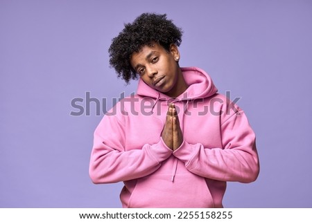 Cunning sneaky African American teen guy holding hands together asking forgiveness, begging please forgive me, hoping or praying looking sly isolated on light purple background.