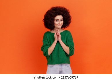 Cunning young adult woman with Afro hairstyle wearing green casual style sweater having tricky plans, looking at camera with cheating face. Indoor studio shot isolated on orange background.