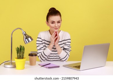 Cunning young adult woman with tricky face scheming and conspiring, pondering devious sly business plan while sitting on workplace. Indoor studio studio shot isolated on yellow background.