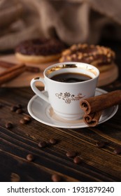 A Cup Of Aromatic Black Coffee, Grains Of Different Varieties On The Table. Morning Espresso Or Americano Coffee For Breakfast In A Beautiful Cup. Still Life. Cinnamon Sticks. Delicious Donut On A Woo