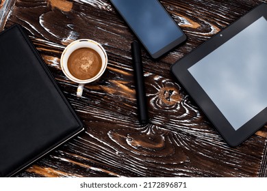 A cup of aromatic coffee, a black tablet, a mobile phone, a notebook with a leather cover and a pen lie on a dark wooden textured surface.