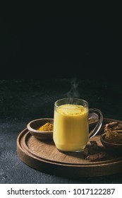 Cup of ayurvedic drink golden milk turmeric latte with curcuma powder on round wooden tray and ingredients above over black texture background. Copy space. Toned image