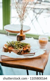 Cup of beautiful cold ice latte and toast with prosciutto and ruccola with wooden vase distichlis spicata on the wooden table. Cafe flatlay situation.