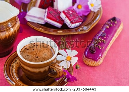A cup of black coffee with an eclair in raspberry glaze on the background of cosmea flowers and sweets