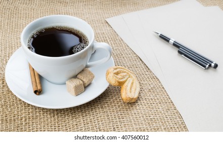 Cup of black coffee with foam, cookie, brown sugar, craft paper, pen and cinnamon stick on sackcloth background - Powered by Shutterstock