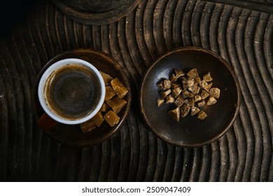 A cup of black coffee on a small plate with a little piece of brown sugar visible from above. A coffee and brown sugar dish with a textured wooden surface in the background. - Powered by Shutterstock