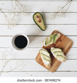 Cup of black coffee, toast with avocado on cutting board, white dried flowers and branches on white wooden table. Overhead view. Flat lay Foto Stok