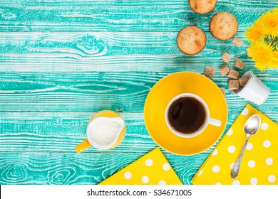 cup of black tea or coffee on yellow plate and yellow milk jug cane sugar, cakes, dandelions, teaspoon on turquoise colored wooden table with yellow napkin at polka dots, top view