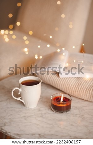 Cup of black tea with paper open book and burning scented candle on marble table over Christmas lights in bedroom. Cozy home atmosphere. 