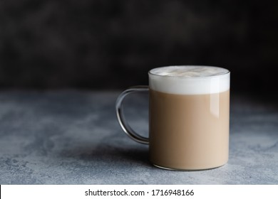 A Cup Of Cafe Latte, Cafe Au Lait Or Chai Latte. The Photo Shows A Beige Or Light Brown Drink With A White Foam On Top. The Drink Is In A Glass Mug, And Is On A Dark Gray Background With Copy Space. 