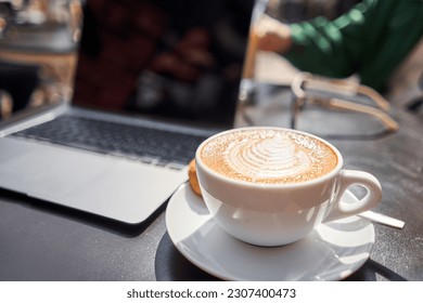 A cup of cappuccino and a laptop on the table in a coffee shop. Workplace in a coffee shop in the morning. coffee break. - Shutterstock ID 2307400473