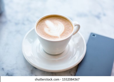 Cup Of Cappuccino With Latte Art On Marbel Cafe Table And Smartphone, Top View.