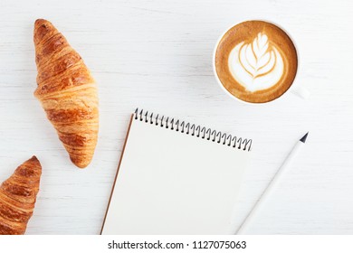 A Cup Of Cappuccino With Latte Art, Two Croissants, Flower And Paper Notebook On White Wooden Table. Top View.