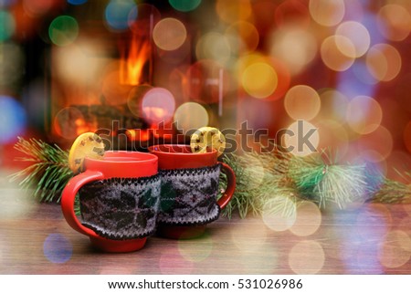 Cup with Christmas ornament near fireplace. Mug in red knitted mitten standing near fireside. Winter holiday concept. Cup of hot drink in front of warm fireplace. Cozy relaxed magical atmosphere.