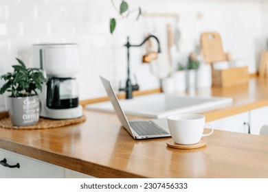 Cup of cofee and laptop on wooden table, view on white kitchen in modern style, drip coffee maker on background - Powered by Shutterstock