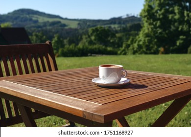 Cup Of Coffe On Wooden Table With Chair.  Blurred Green Trees And Hill Tops Background. Coffee Break In Countryside. Picnic, Garden Party Snack, Relaxation Concept. No People, Selective Focus.