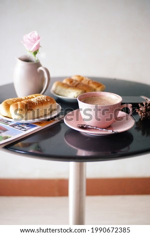 A cup of coffee, 2 plates of bread, a magazine, a faux rose and a handphone on the round table. View of morning breakfast. Selective focus.