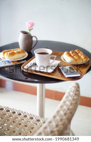 A cup of coffee, 2 plates of bread, a phone, a magazine, a faux rose in a vase and a wooden tray on the round table. A set of table and a white chair. Selective focus
