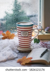 A cup of coffee against window view in a rainy day with raindrop on a glass and cozy atmosphere with red orange autumn leaves, books and pen and bokeh of the light in background